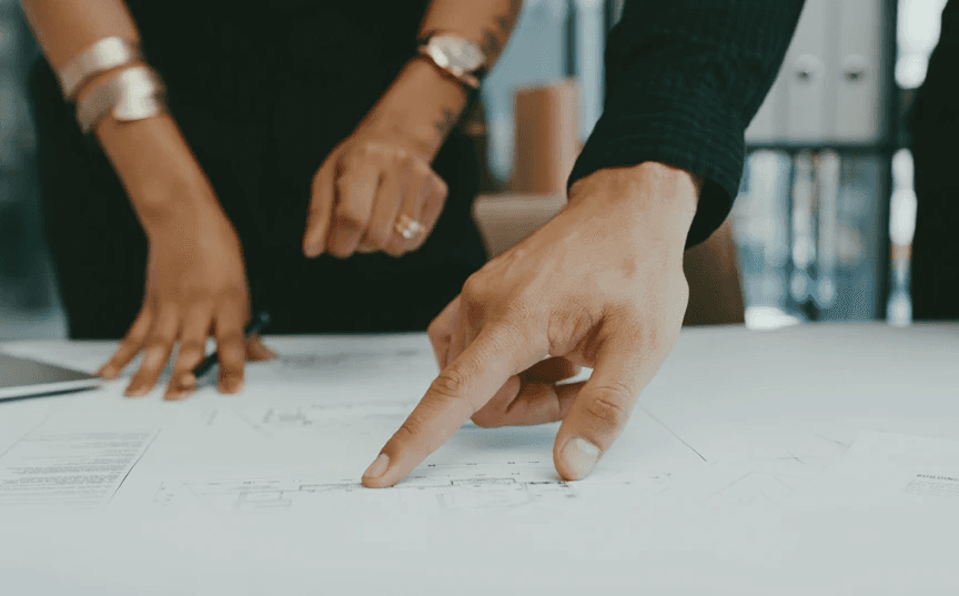 Hands collaborating over service design maps and architectural floor plans, representing embedded partnership in discovery, design, delivery, and optimization.