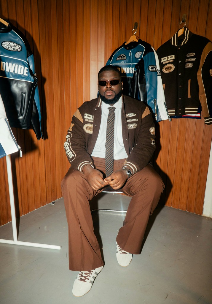 Man in letterman jacket and brown pants sits.