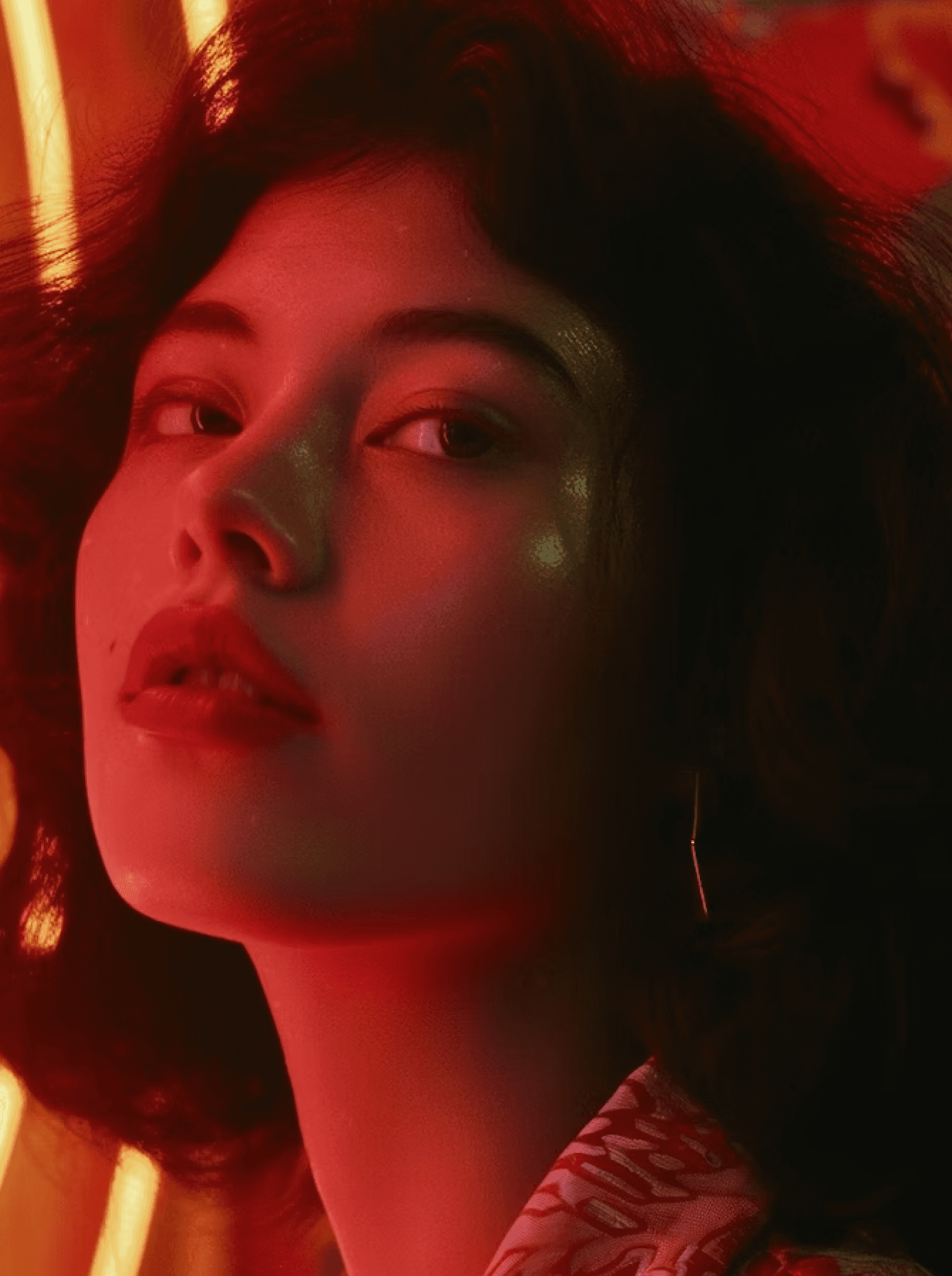 A person with wavy hair is illuminated by vibrant red neon lights, creating a dramatic and colorful portrait against a blurred background.