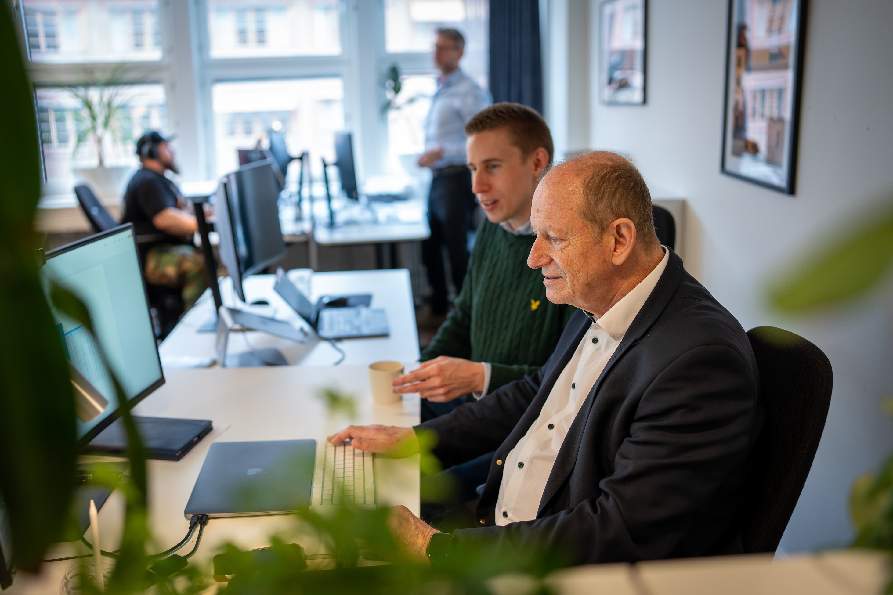 A young person with curly hair smiles at the camera while sitting in front of a laptop against a dark background.
