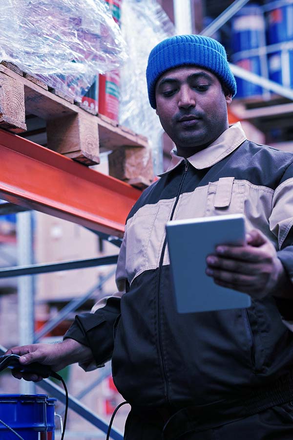 Close-up of a man looking at an electronic tablet in a warehouse setting, with blurred shelves and boxes visible in the background.