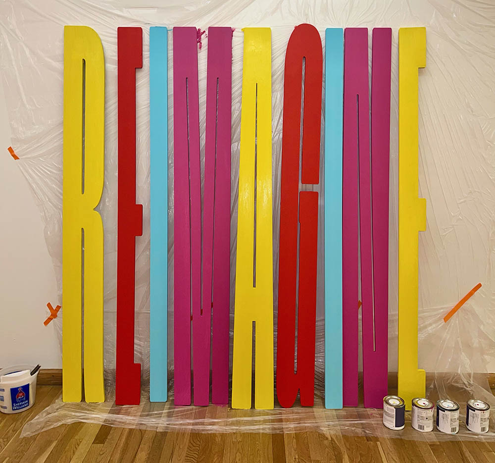 A yellow, red, cyan and magenta painted 3d text that says: REIMAGINE.