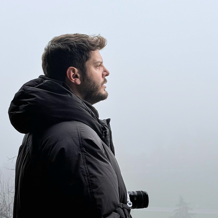 A side-profile of a young person with dark, wavy hair, wearing silver sports sunglasses and a black sweater, sitting in front of a blurred teal couch.
