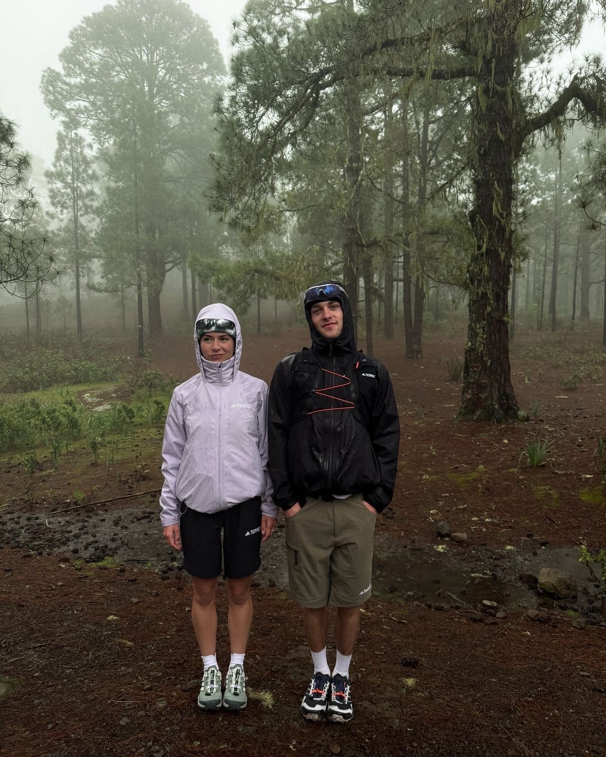 A smiling woman with her arms crossed, standing against a dark green background. She has long, dark hair.