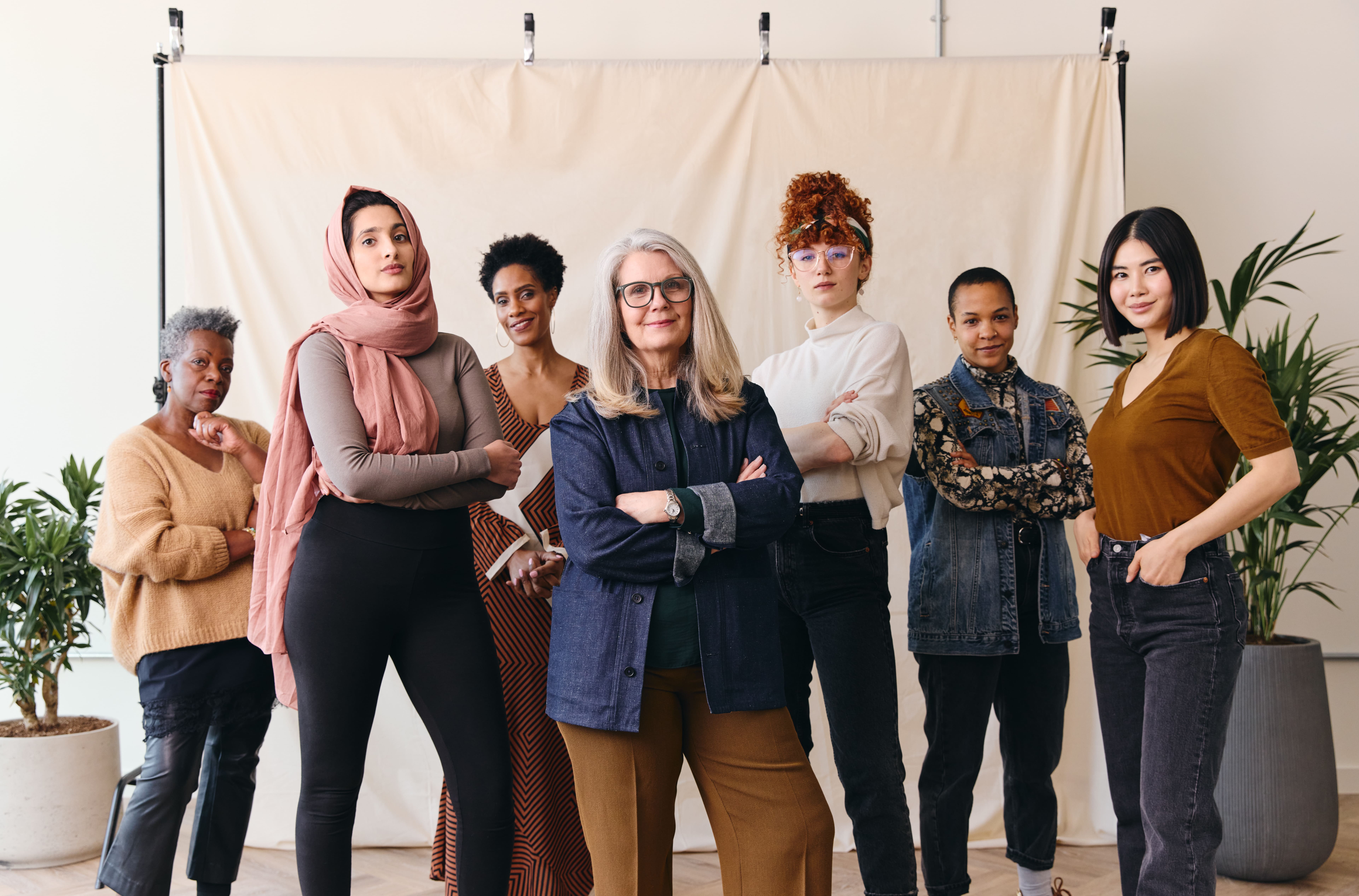 Imagem de um grupo diverso de mulheres de diferentes idades, etnias e estilos, todas com expressões confiantes. Elas estão posicionadas em frente a um fundo neutro, transmitindo uma mensagem de empoderamento, diversidade e inclusão. Cada uma delas exibe sua individualidade através de roupas e acessórios distintos, reforçando o sentimento de união na diversidade. A composição visual sugere força e solidariedade, celebrando a representatividade feminina em um espaço acolhedor e moderno, com plantas ao fundo que complementam a ambientação.