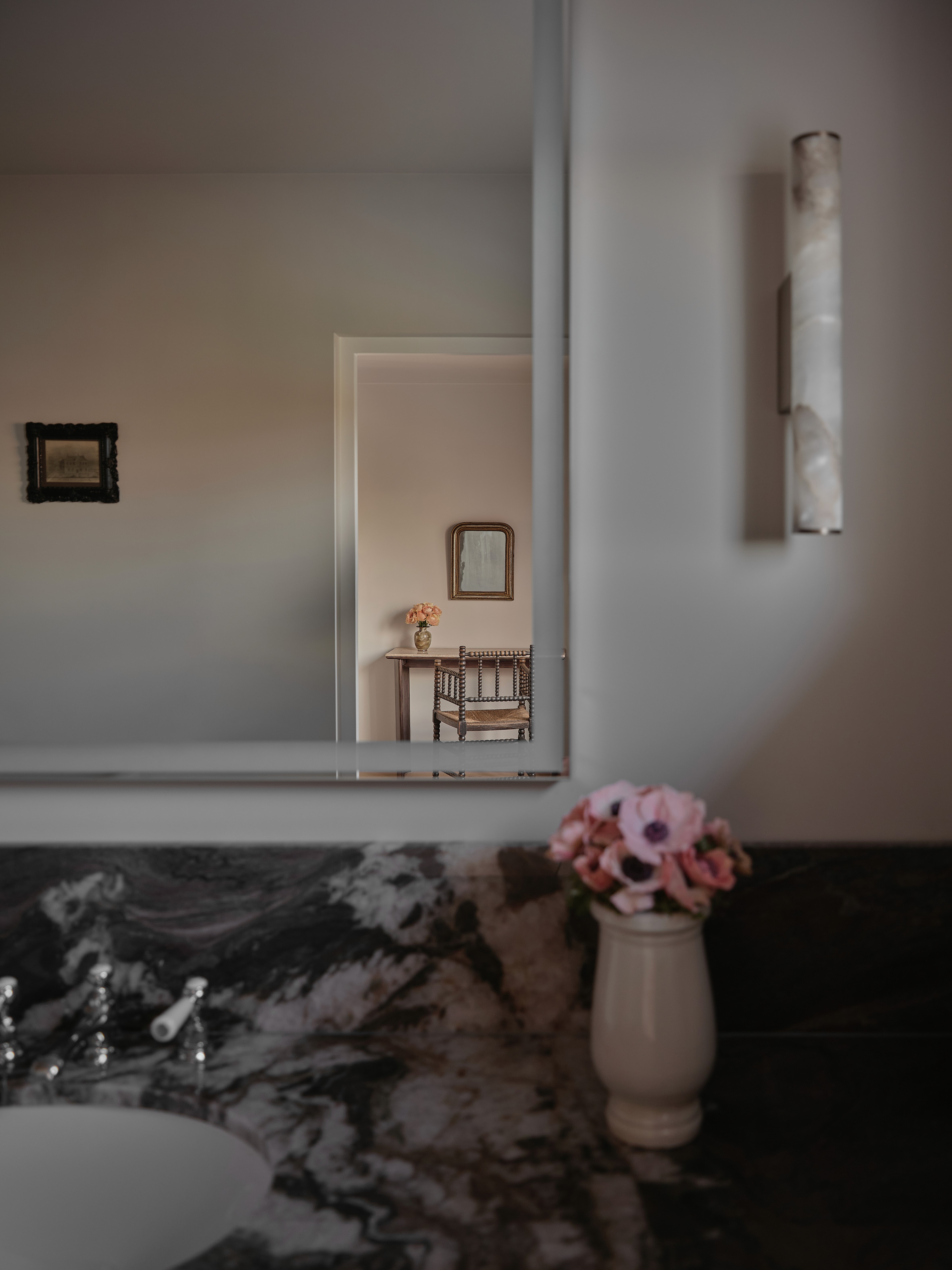 Bathroom vanity with dramatic veined marble countertop, alabaster wall sconce, pink anemones in ceramic vase, and mirror reflecting bedroom