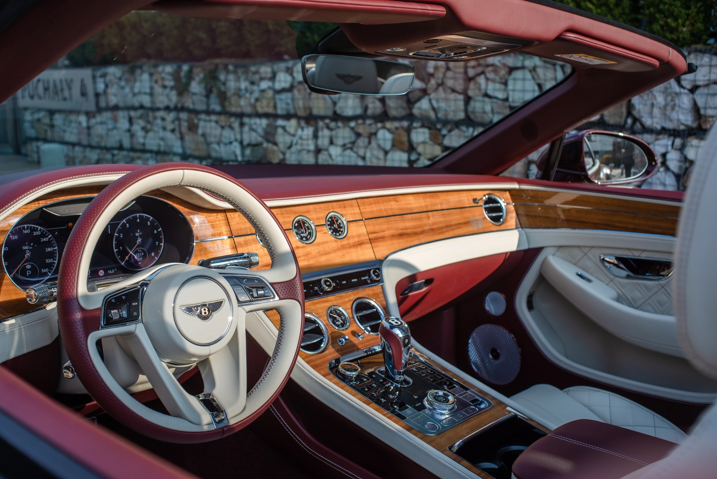 Wnętrze luksusowego samochodu Bentley z drewnianymi elementami i skórzaną kierownicą. Czerwone akcenty i białe siedzenia.