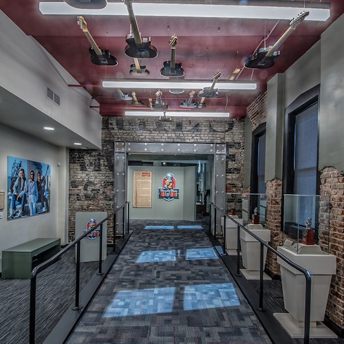 Muzejní chodba s pověšenými kytarami, vitrínami, odhalenými cihlovými stěnami a nástěnnou malbou, na které jsou viditelní sedící návštěvníci.