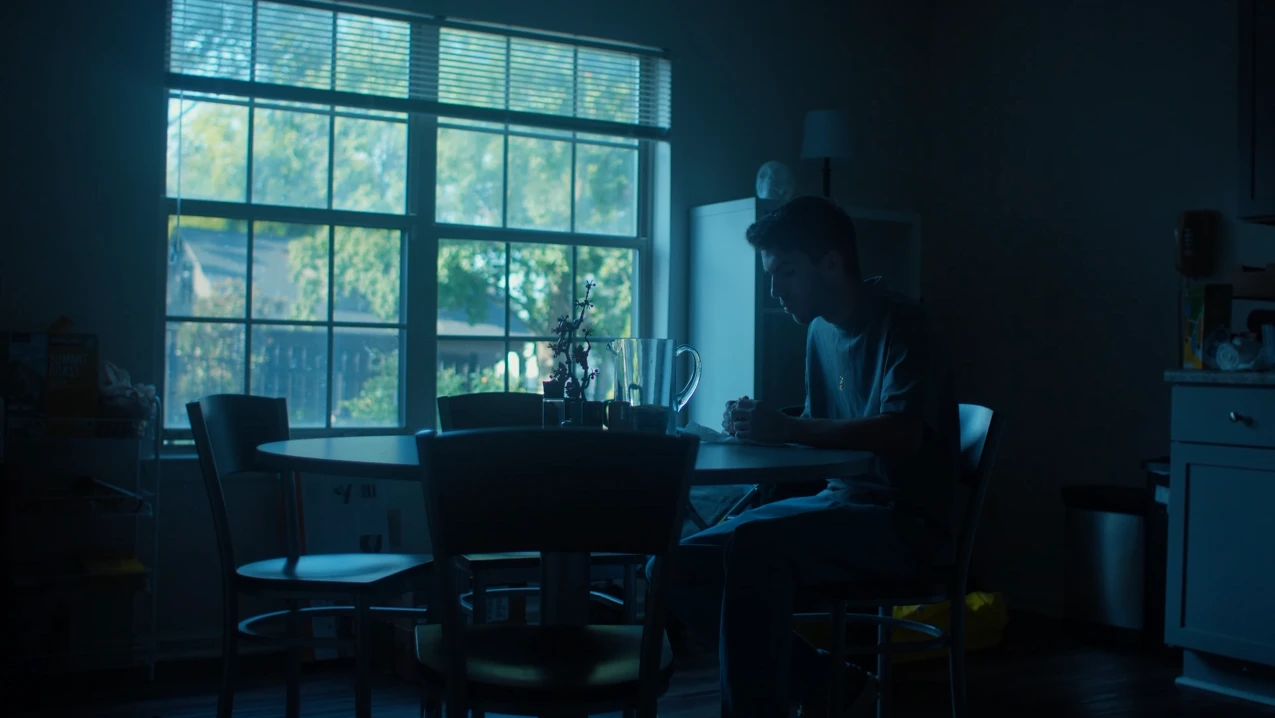 Un jeune homme est assis seul à une table ronde dans une pièce sombre, éclairée par la lumière bleue d'une fenêtre.