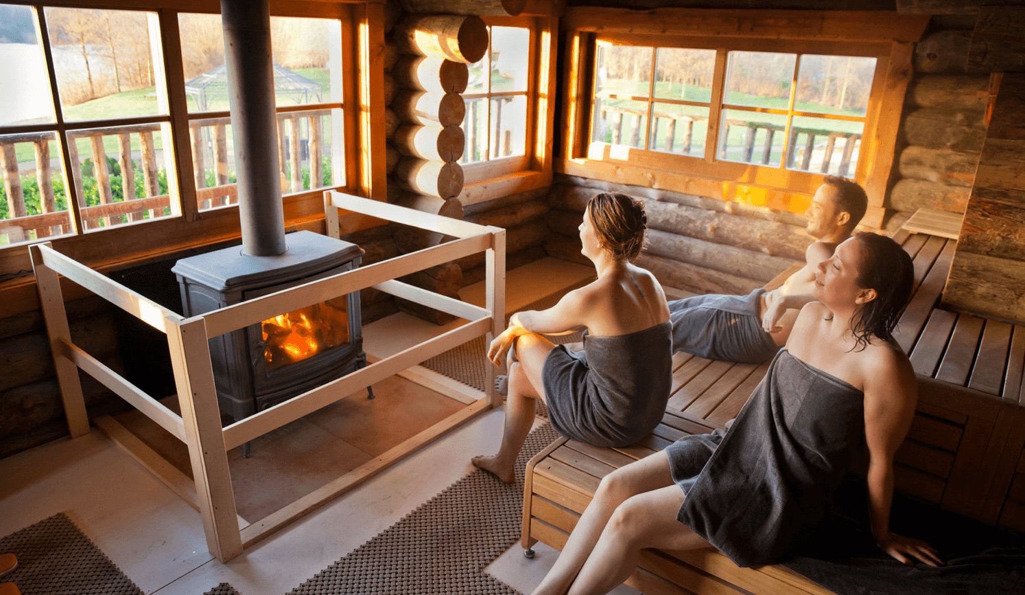 Interieur van een wellnessruimte met houten wanden en een persoon in een handdoek die ontspant in een sauna. Er is een groot raam met uitzicht op de natuur.