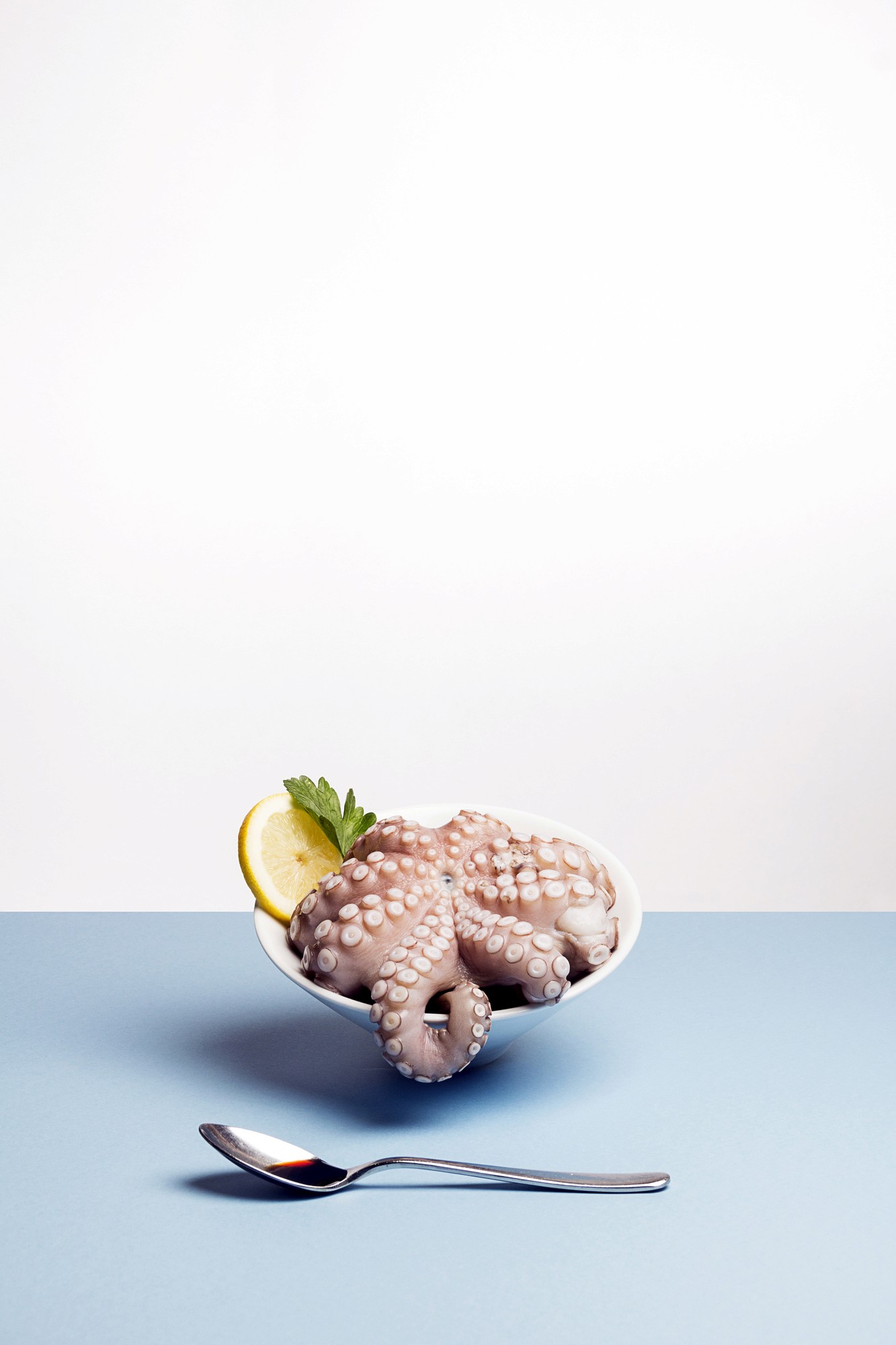 Photo studio sur une table au fond bleu ciel d'un poulpe entier dans un bol avec une tranche de citron vert et d'une cuillère disposée au premier plan contenant de la sauce soja