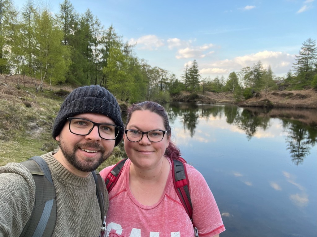 A selfie of Martin and April stood in front of the Holme Fell reservoir. The water reflects the trees behind and the blue sky above.