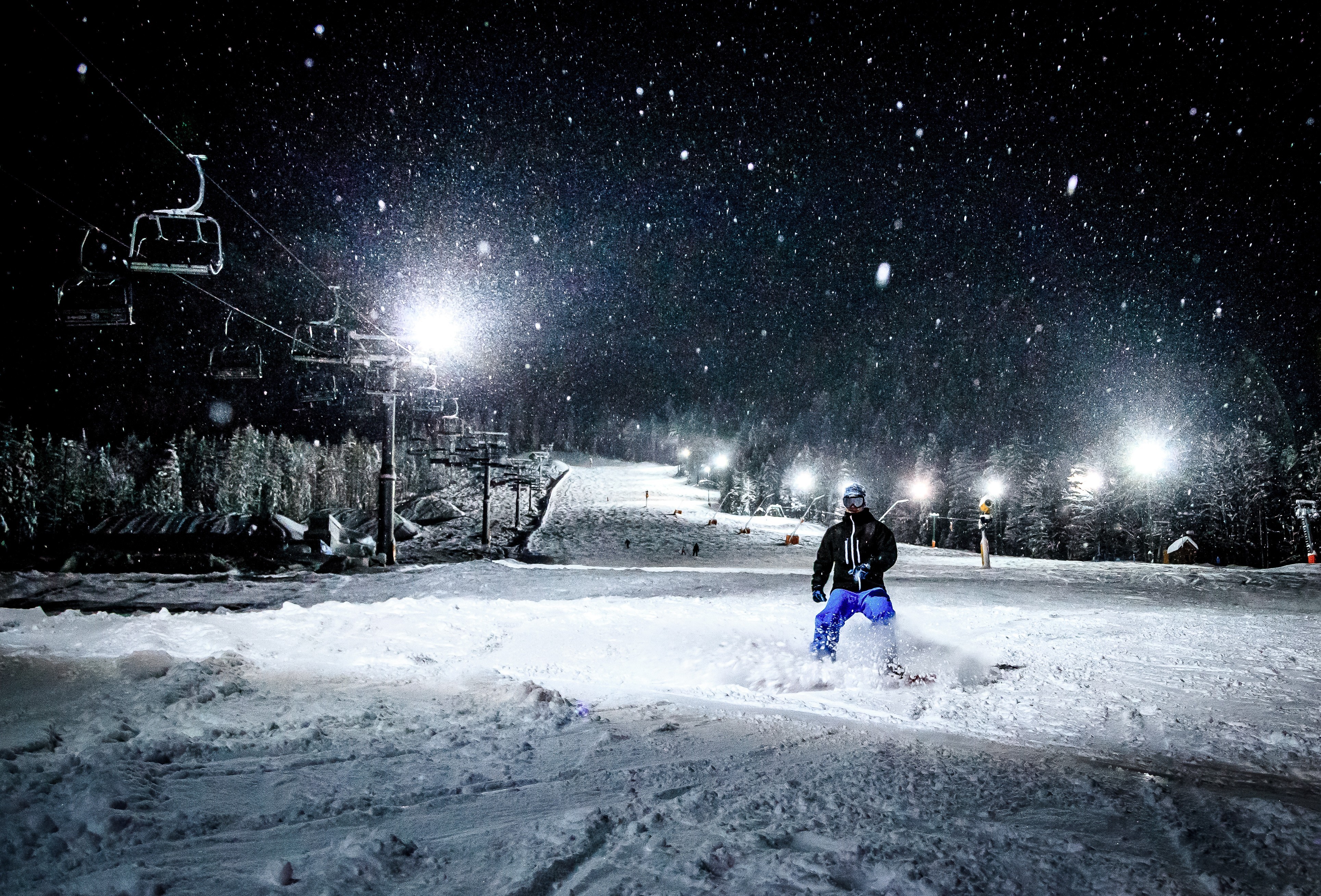 A snowboarder rides down a floodlit slope at night as snow falls and chairlifts hang overhead.