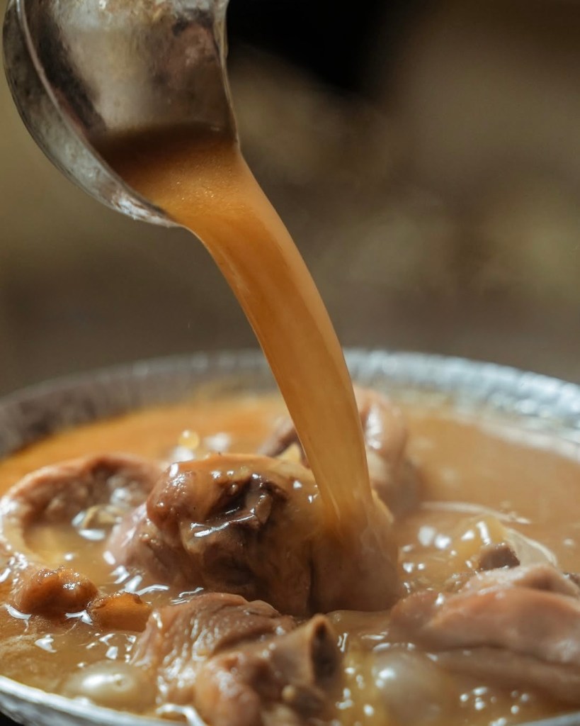 A close-up view of a ladle pouring rich, brown sauce over tender pieces of chicken in a clay pot, showcasing a traditional Taiwanese dish.