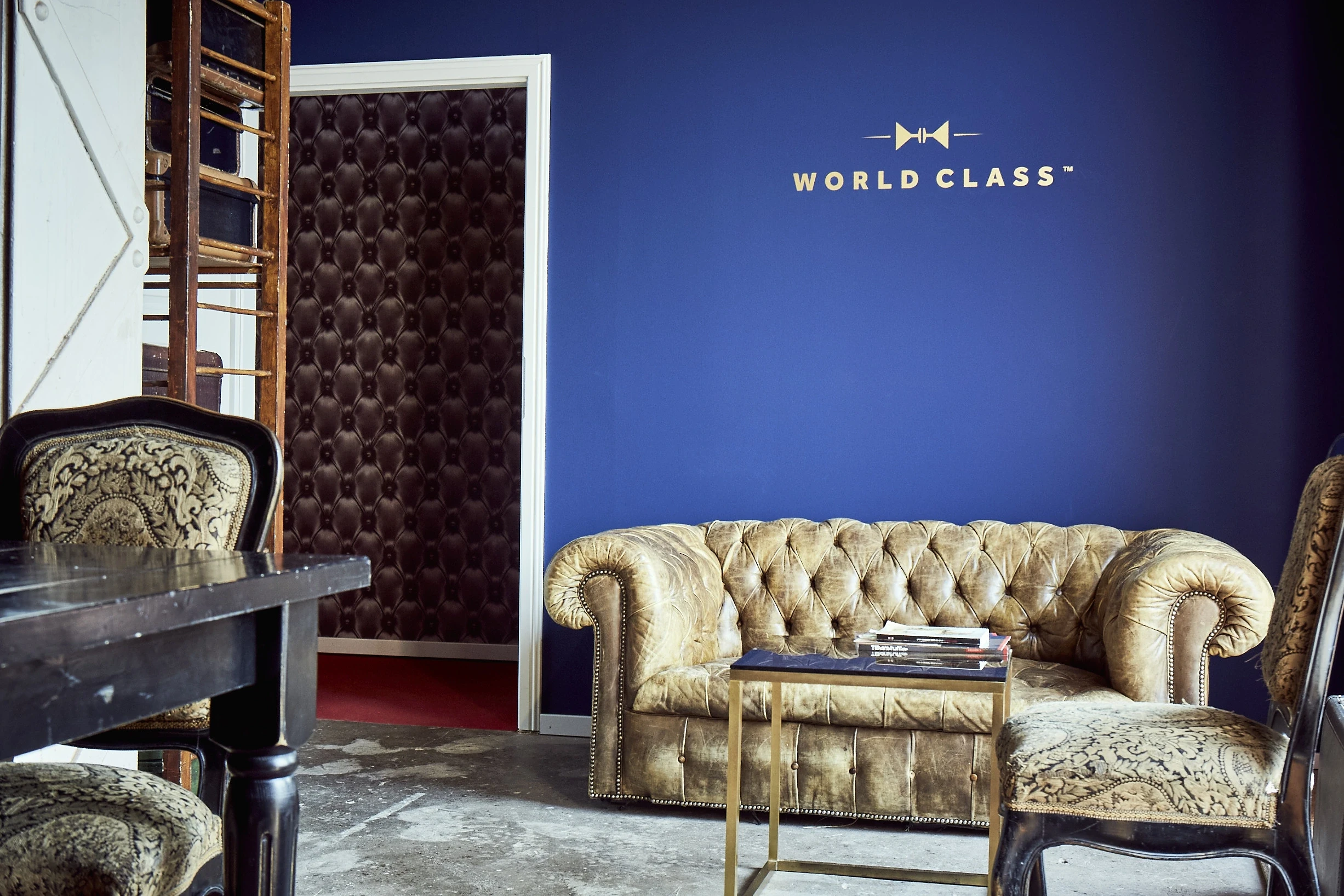 World Class lounge at DIAGEO BCB17 with a golden Chesterfield sofa against a deep blue wall with golden brand logo and a patterned dark doorway.