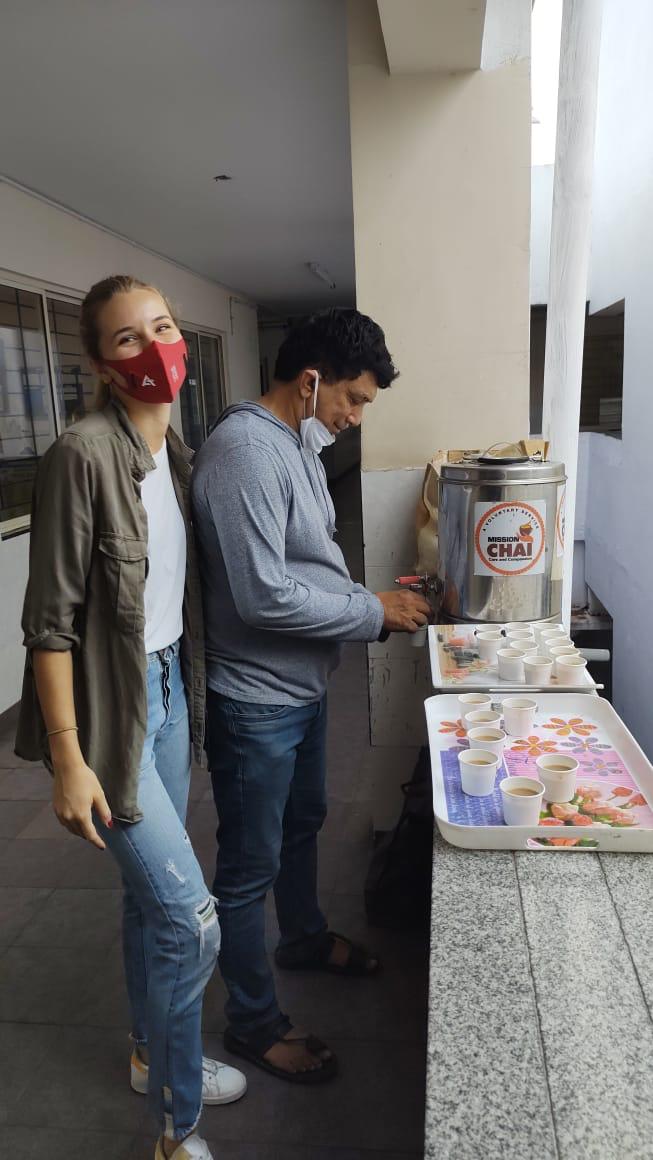 Two volunteers serve tea from a "Mission Chai" dispenser into paper cups on trays, smiling in a hospital corridor.