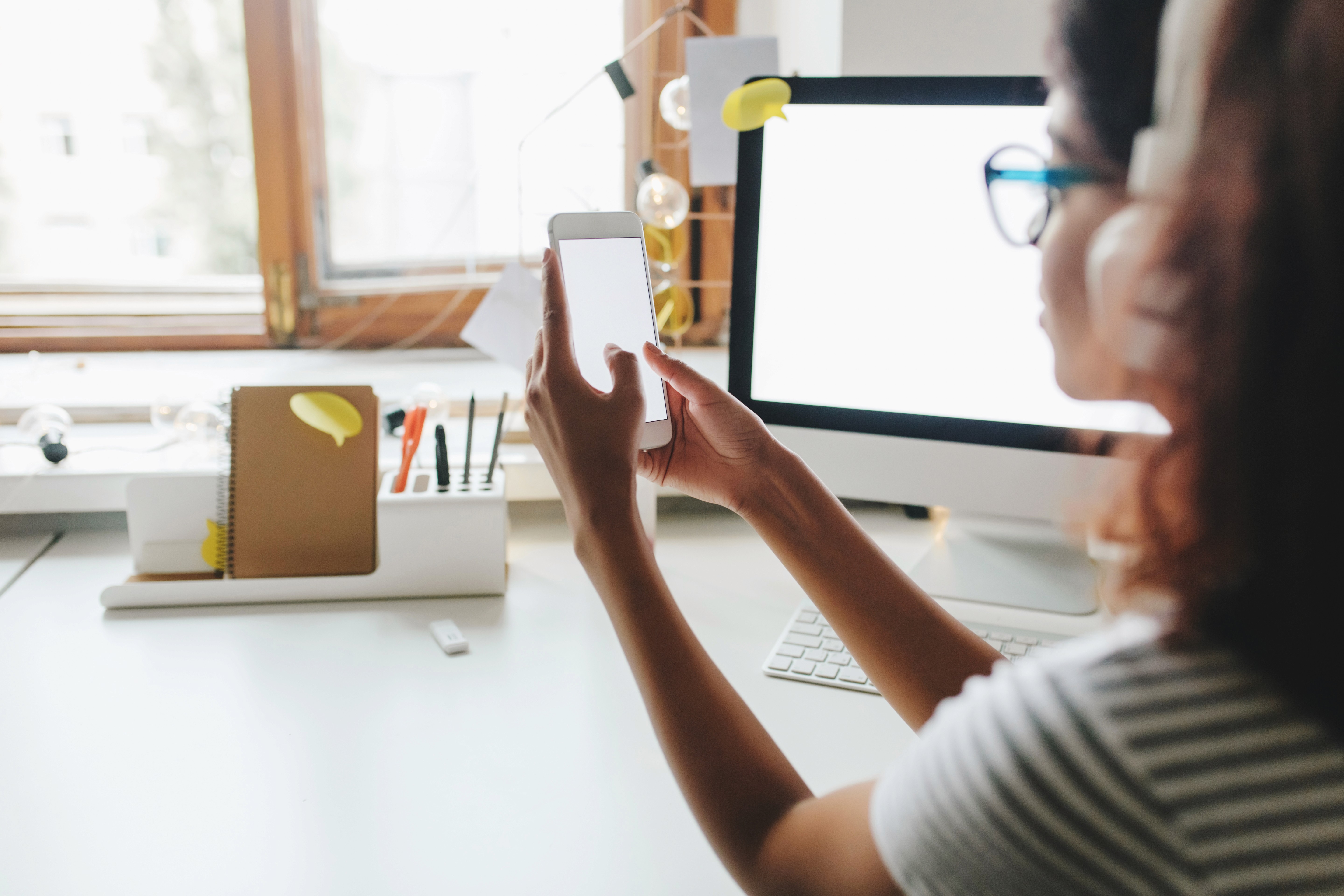 Woman wearing headphones uses a smartphone at her desk beside a desktop computer, likely reviewing or testing a mobile experience.
