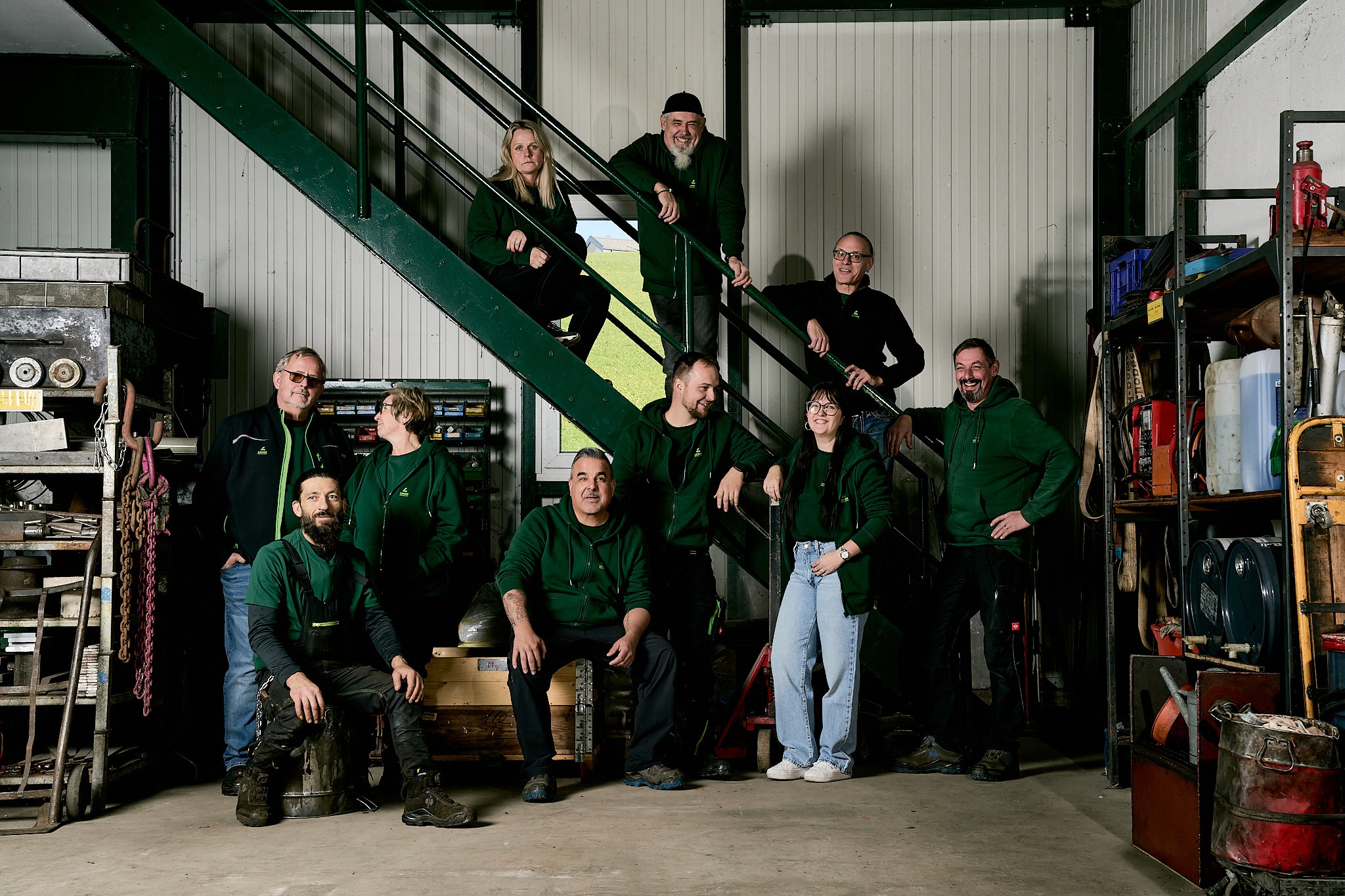 Gruppe von 11 Personen in grünen Oberteilen, die in einem Werkstattgebäude posieren, einige stehen auf einer Treppe.