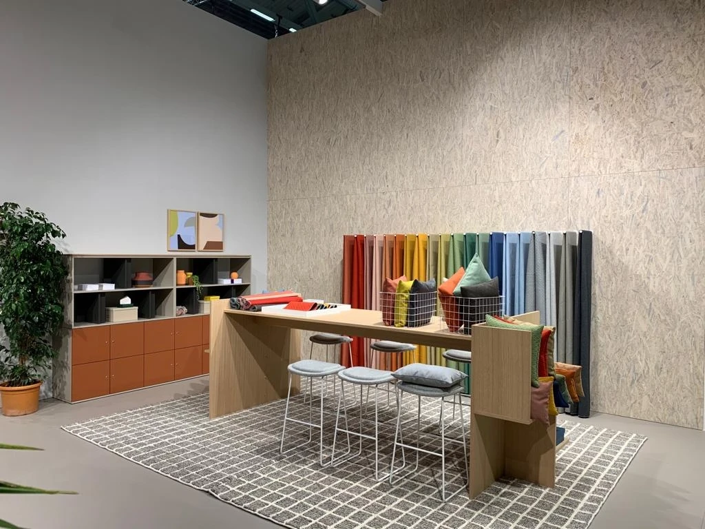 Work area at the HAWORTH ORGATEC stand with a wooden high table, white stools, a colorful row of upright felt panels and an orange sideboard on OSB walls.