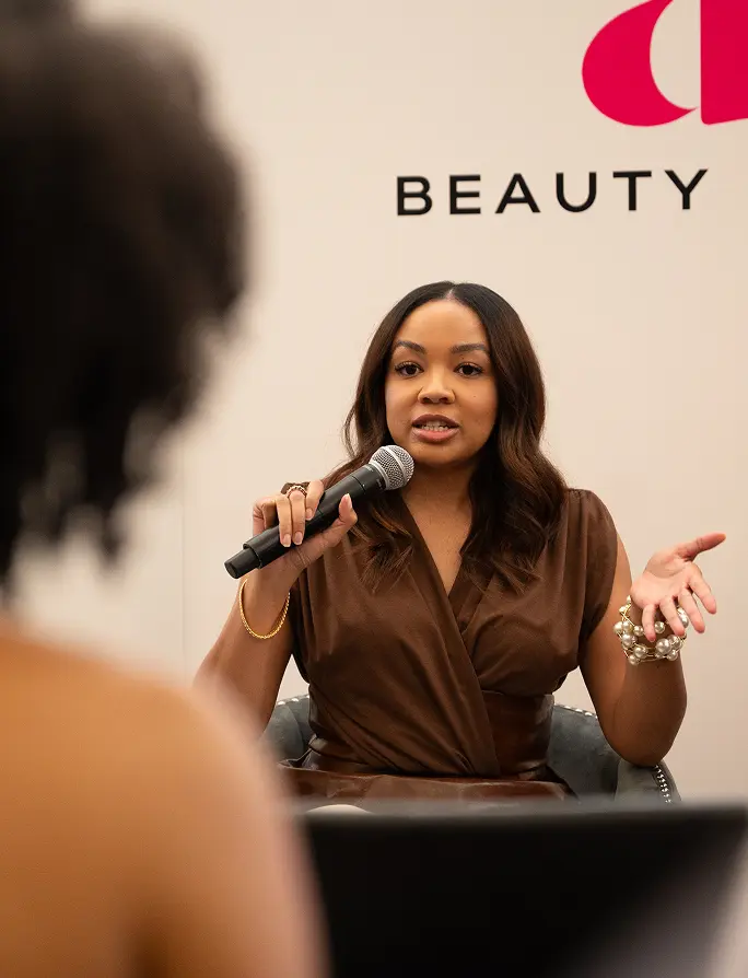 A woman in a brown outfit confidently speaks into a microphone at a beauty event. The word "BEAUTY" is partially visible on the backdrop.