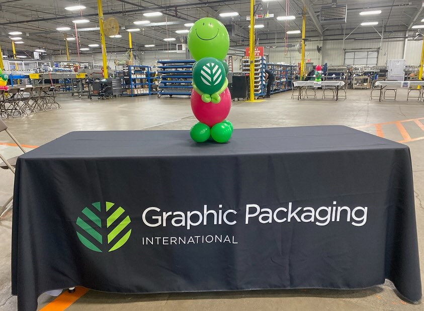 A colorful green and pink balloon sculpture shaped like a smiley character is placed on a black tablecloth with the "Graphic Packaging International" logo, set in a spacious industrial warehouse with shelves and equipment in the background.