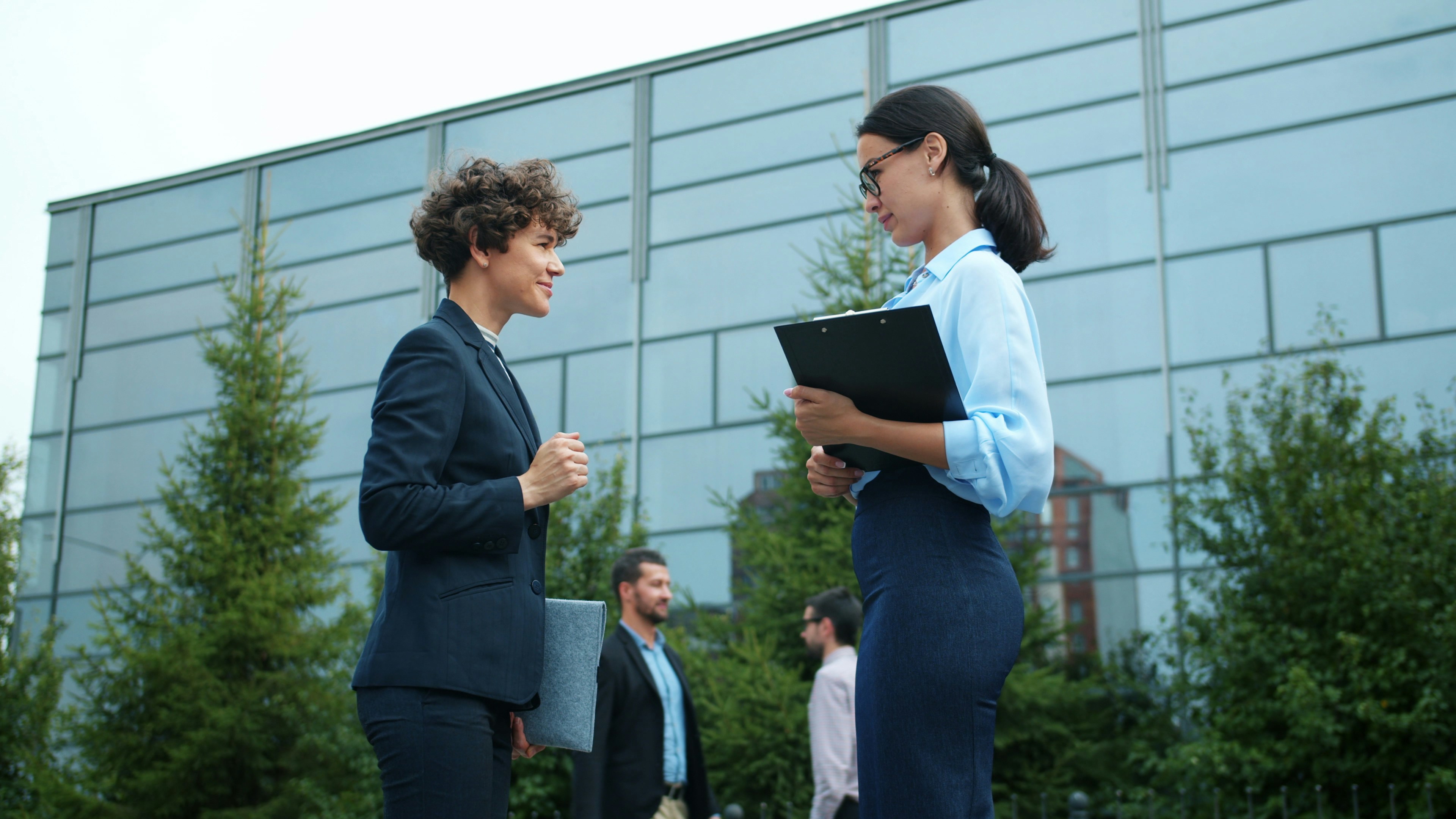 Two businesswomen converse outside modern office building.