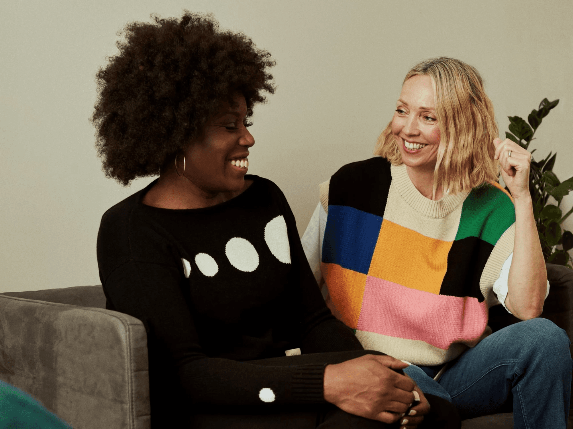 Two women sit together on a grey couch, sharing a joyful conversation. The woman on the left has voluminous dark curly hair and wears a black sweater with white circular patterns, while the woman on the right has blonde wavy hair and wears a vibrant, multi-colored patchwork sweater vest over a white shirt. They are both smiling and looking toward each other in a relaxed, indoor setting with a green plant visible in the background.