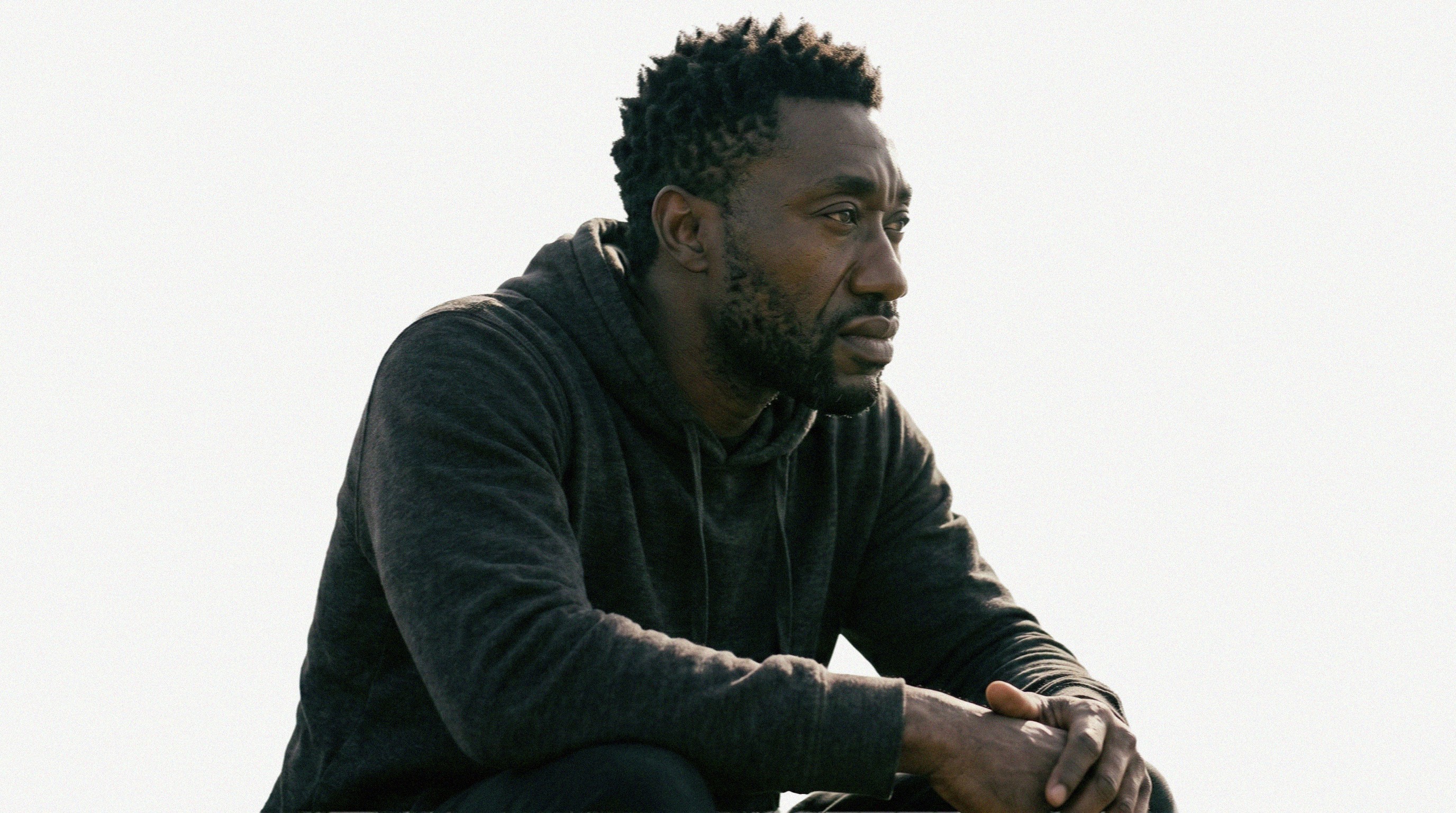 A person in a dark hoodie sits outdoors with a distressed expression, hand on forehead, conveying a sense of contemplation or stress against a plain background.