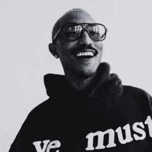 Pharrell, a smiling man wearing glasses and a black hoodie with the text "we must" displayed prominently.