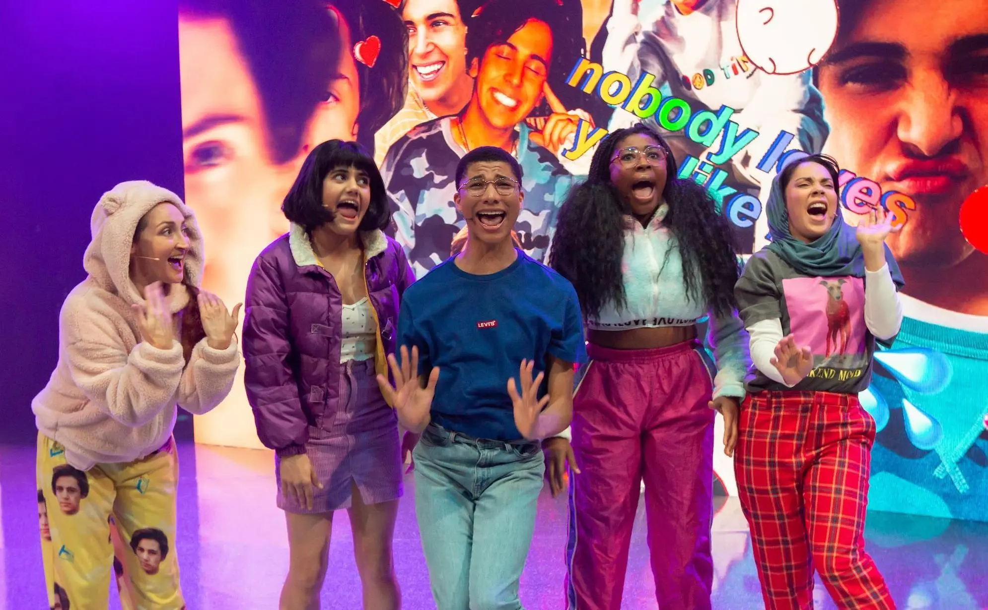 A group of five excited young actors stand in bright costumes, yelling joyfully in front of an oversized digital collage of a teen idol’s face and colourful text, suggesting an explosive pop-fan energy.