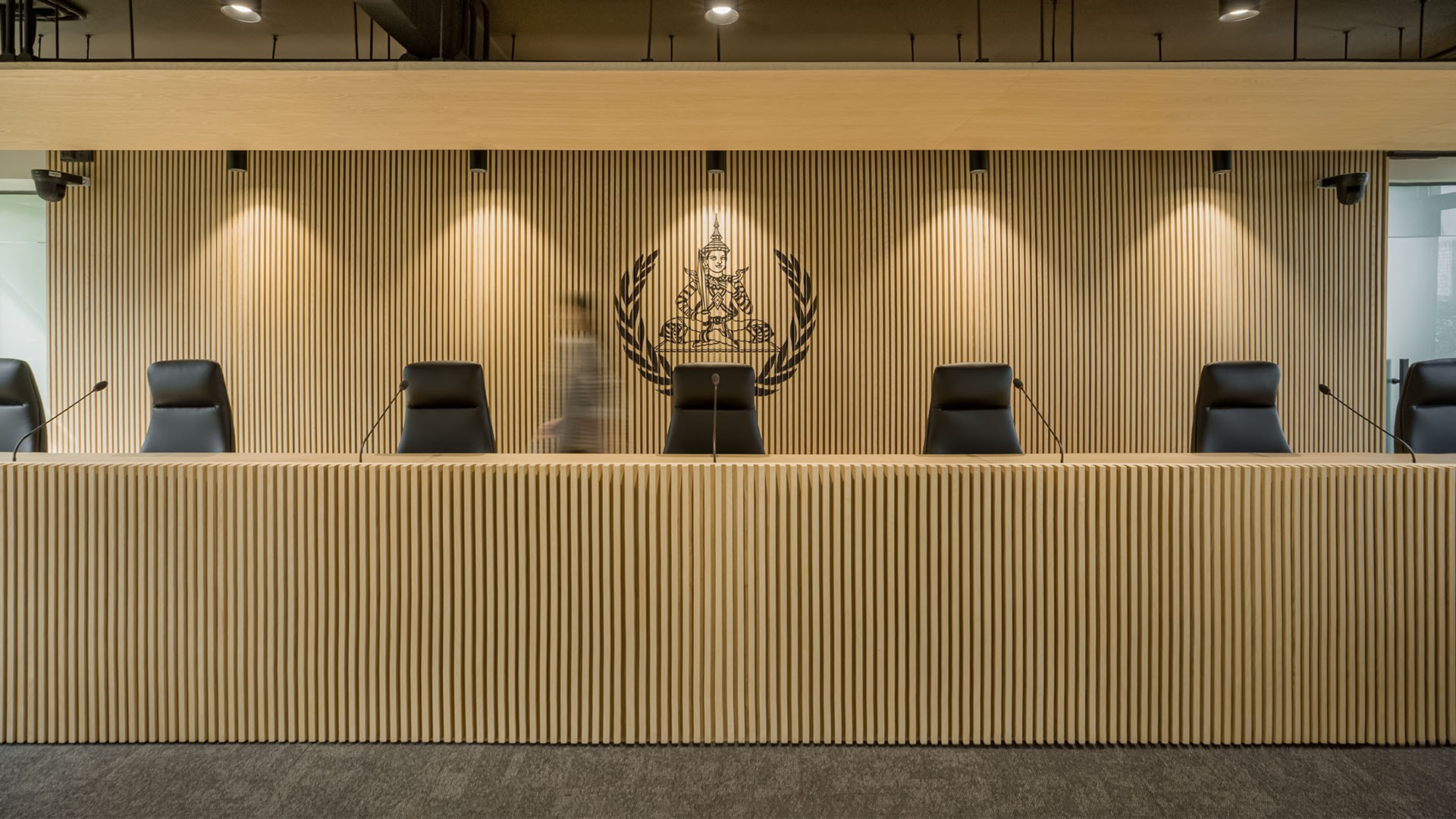 The ECCC Courtroom in Phnom Penh, featuring a light wooden slatted wall with a prominent Cambodian crest, and a row of black upholstered chairs for the judges' seating area.