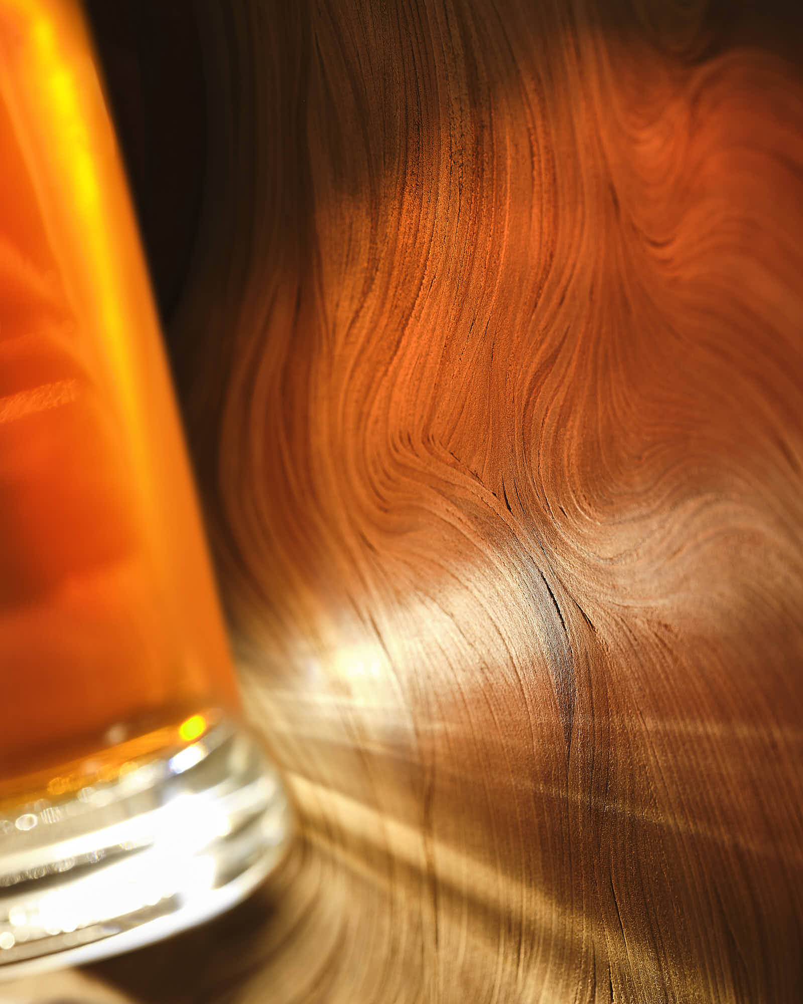 A glass of amber-colored liquid sits on a wooden surface with warm lighting reflecting off it.