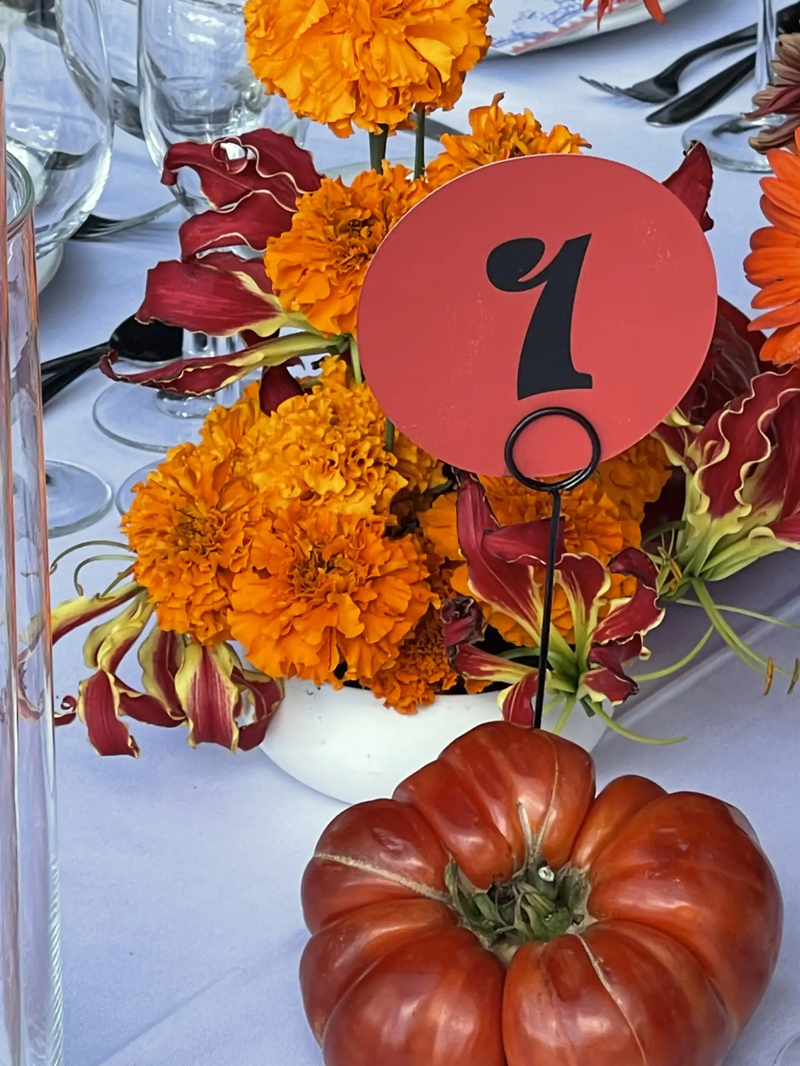 Detalle de centro floral naranja con número de mesa y gran tomate decorativo en evento con estilo orgánico.