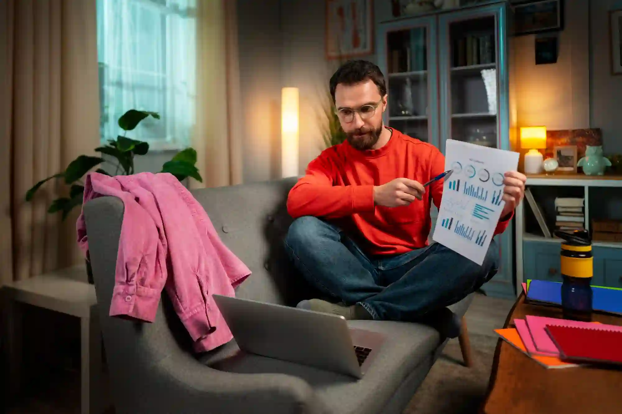 Man sitting on a sofa pointing to a chart on paper while talking on a video call on his laptop.