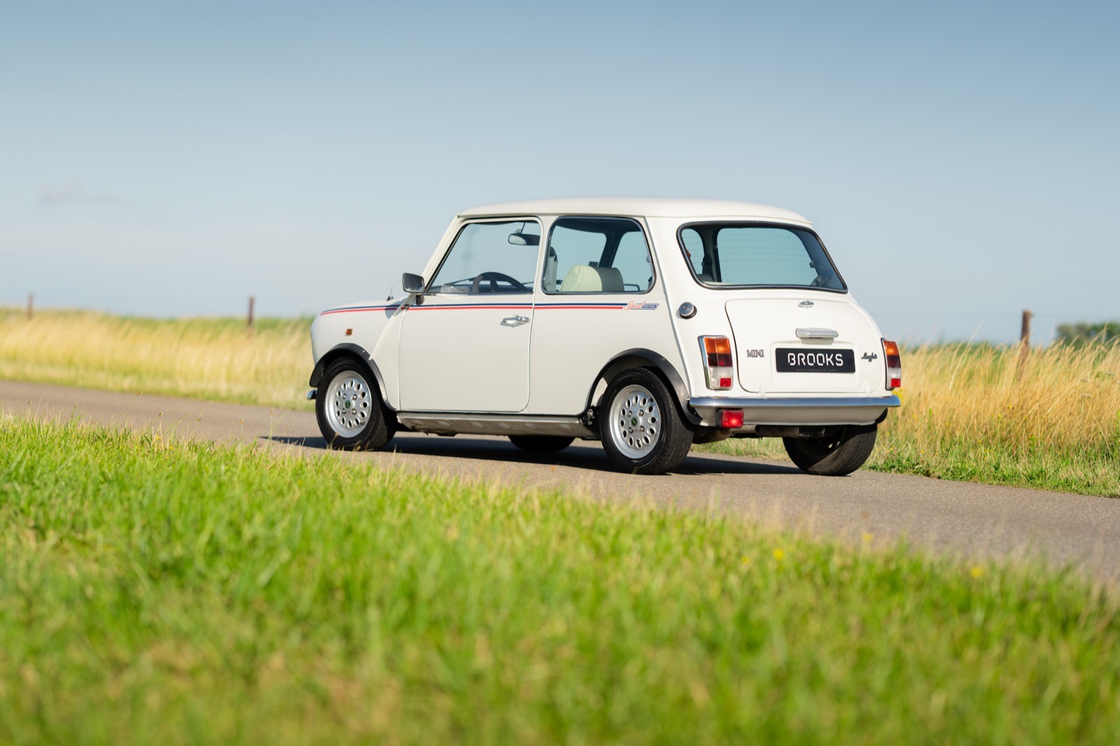 A classic white car parked on a rural road, surrounded by green grass and fields under a clear blue sky.