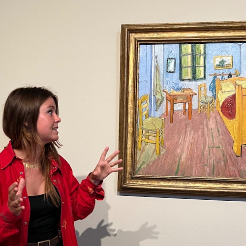 一位穿着红色衬衫的女士热情地比划着一幅挂在墙上的画，这幅画描绘了一间有椅子、桌子和床的房间。