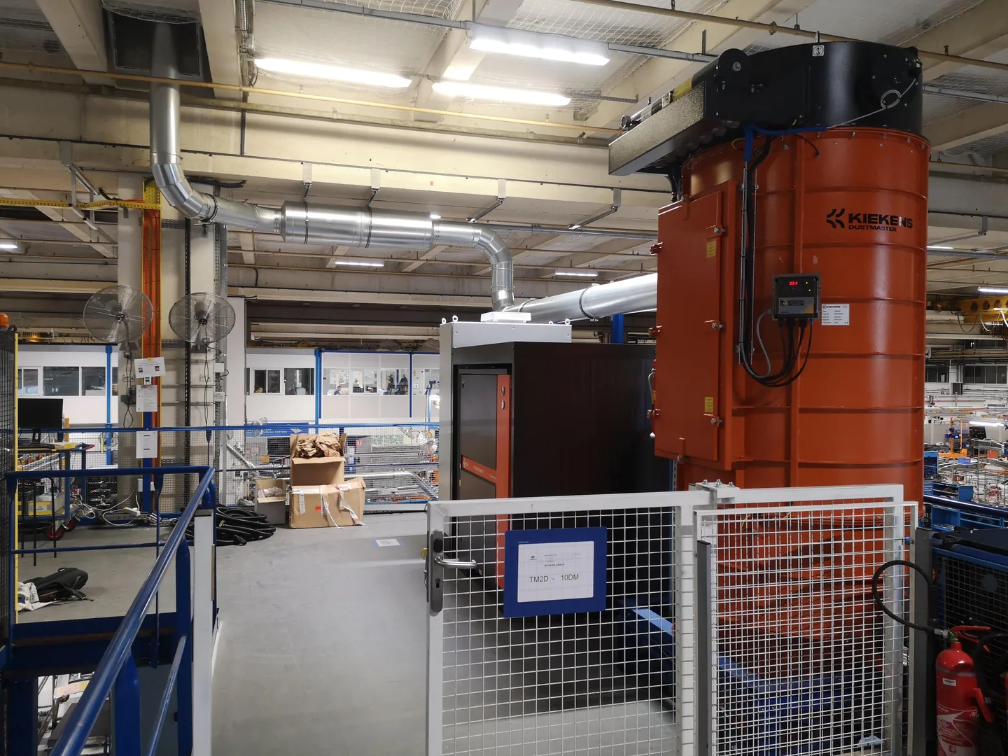 Cyclone d’aspiration orange protégé par une enceinte grillagée, avec tuyauteries et équipements de filtration dans l’atelier.