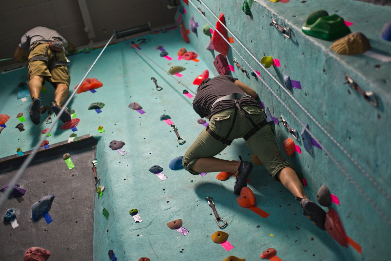 Two climbers on a teal rope wall, one halfway up and one near the top anchors