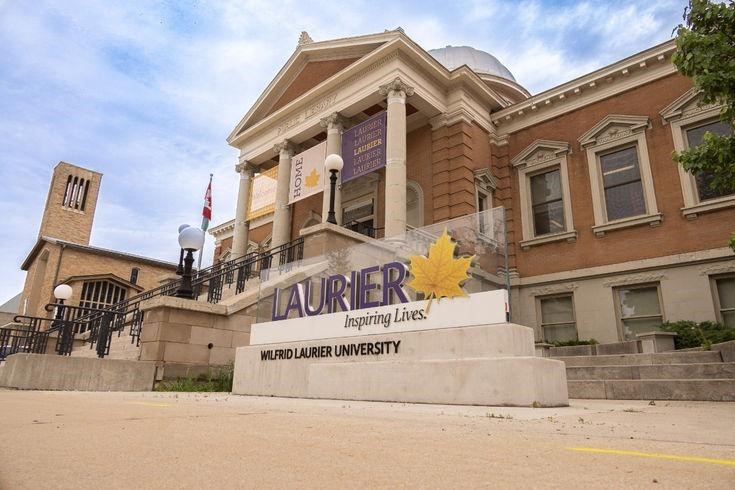 This image shows the front of Wilfrid Laurier University, specifically one of its iconic campus buildings. The sign prominently displays the university's branding with the slogan "Inspiring Lives" and a golden maple leaf symbol. The architectural style features classical elements like columns and a dome, emphasizing the institution's historic and academic presence. Wilfrid Laurier University is located in Ontario, Canada, with main campuses in Waterloo and Brantford.