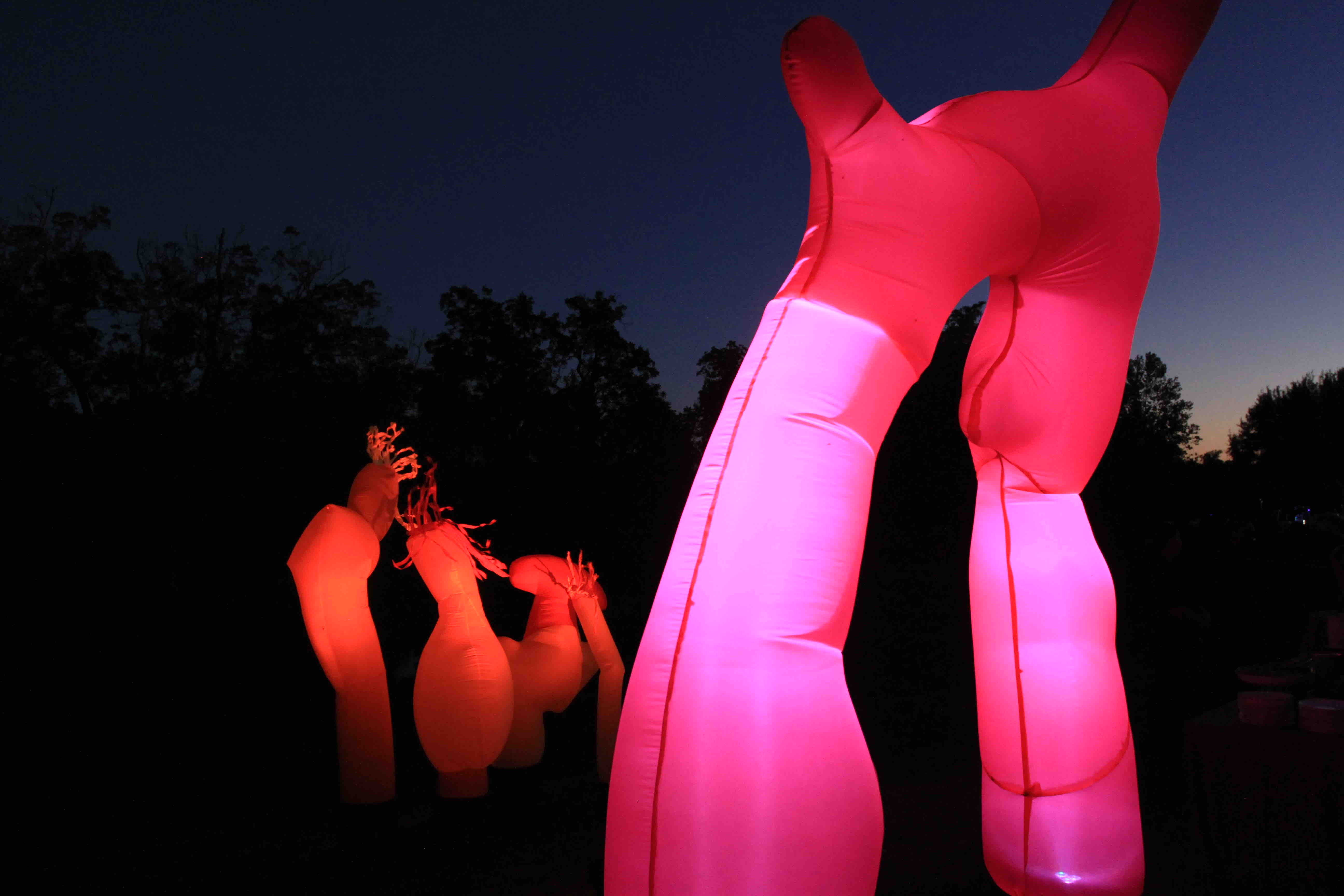 Two hands raised in the air, illuminated in pink, against a twilight sky with silhouettes in the background.