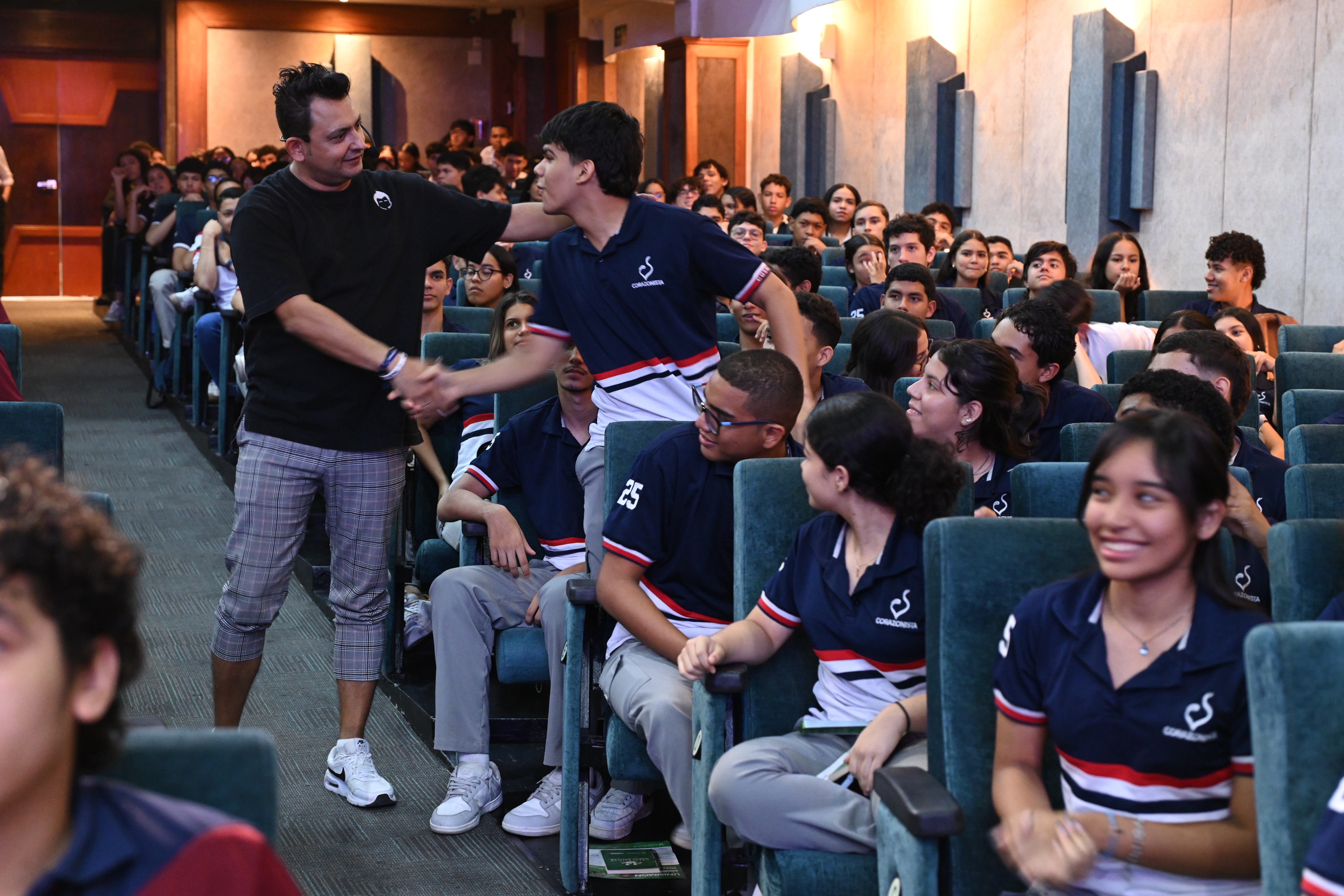 Jorge Baena caminando entre el público de un auditorio, estrechando la mano de un estudiante y conversando con jóvenes uniformados