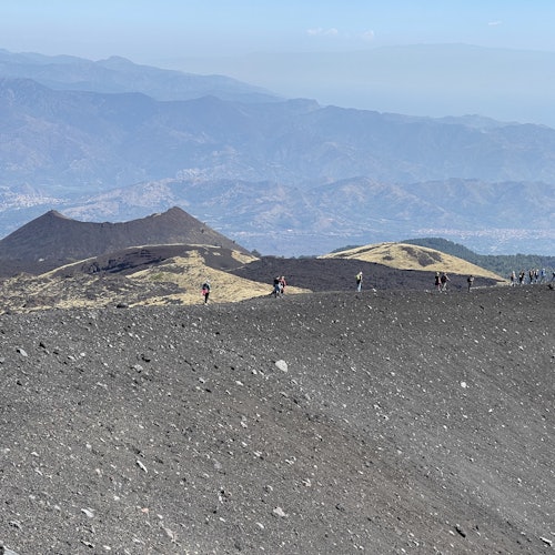 Pessoas a caminhar por uma paisagem rochosa e vulcânica, com cadeias montanhosas e um céu enevoado ao fundo.
