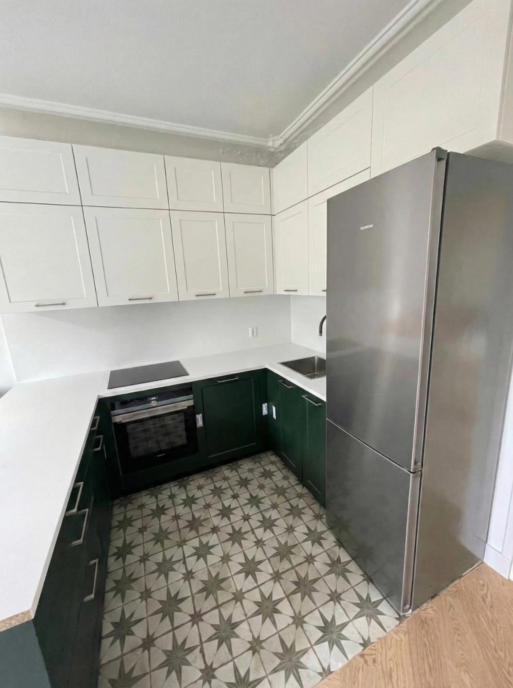 Cuisine équipée avec meubles bas vert forêt, placards hauts blancs et sol en carreaux de ciment à motifs gris.