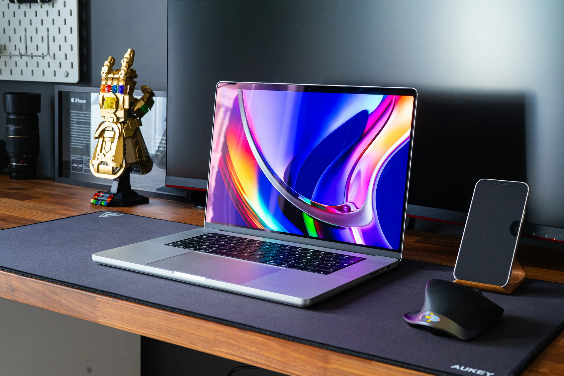 A laptop is sitting on a wooden desk with a large, colorful abstract image displayed on the screen, a phone standing upright, a mouse, and a gold superhero gauntlet figurine nearby.