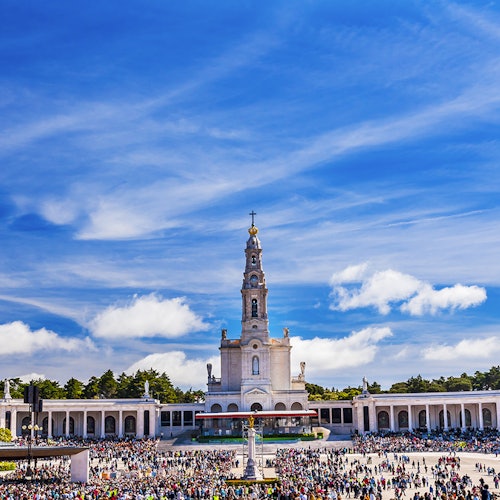 A large crowd gathers in front of a grand church with a tall bell tower under a bright blue sky with scattered clouds.