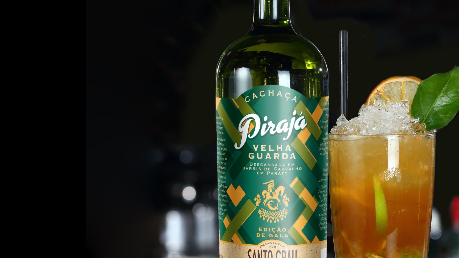 A bottle of Pirajá cachaça with a green and gold label sits beside a cocktail in a glass with ice, lime, and an orange slice garnish. The background is dark.