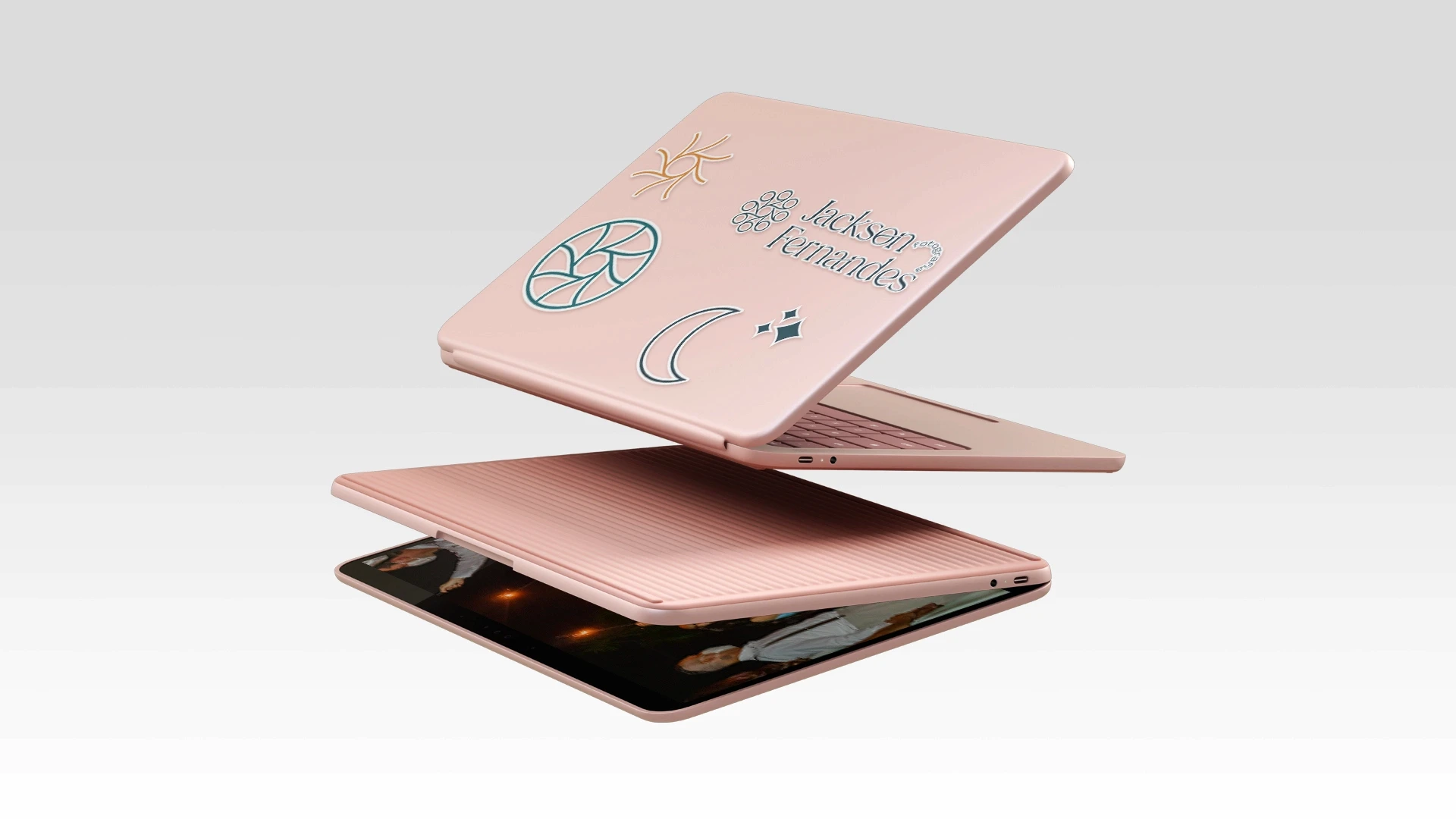 Dispositivo do tipo notebook/tablet na cor rosa em ângulo flutuante, com a parte traseira da tampa personalizada com adesivos ilustrativos de sol, lua, obturador e a marca Jackson Fernandes.