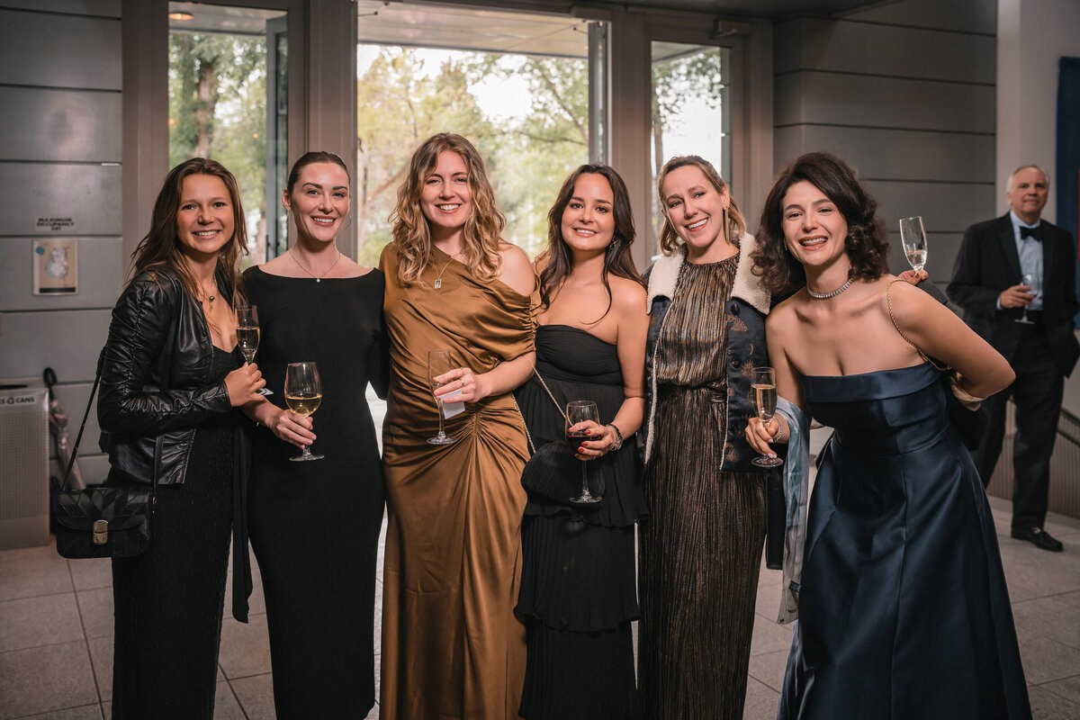 Six smiling guests in evening dresses holding drinks at an indoor reception