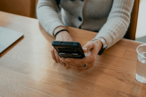 A person’s hand with a smartphone sits on a wooden table, with a glass and a notebook nearby.