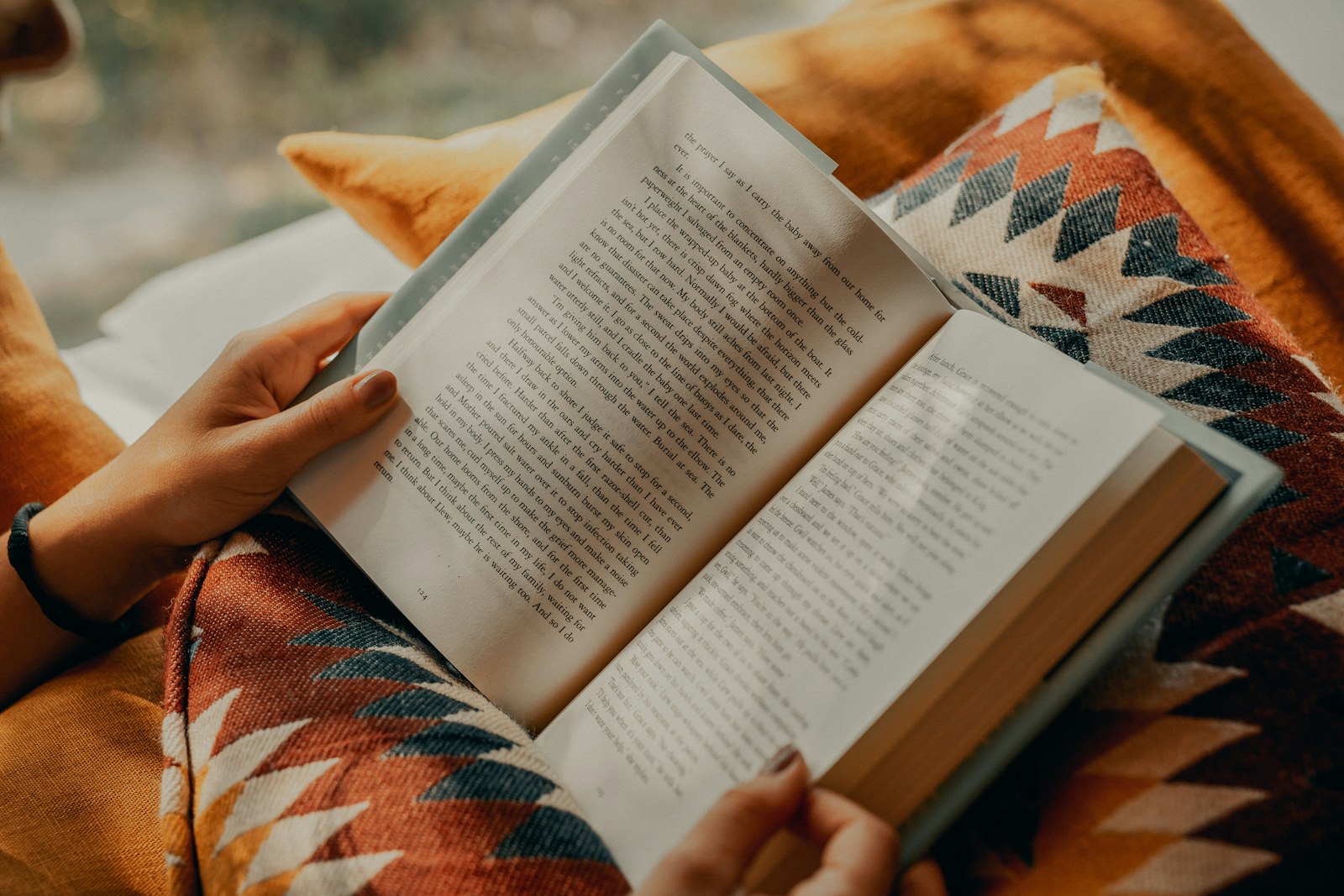 Hands holding an open book over a warm orange patterned cushion.
